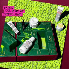 A festive Kiehl’s Advent Calendar box is opened to reveal numbered green drawers with graffiti-style neon yellow and black designsinside. Several skincare products are displayed in front, including the Calendula Herbal-Extract Toner, Creamy Eye Treatment with Avocado,and Midnight Recovery Omega-Rich Cloud Cream. A bright pink banner at the top left reads: “UNBOX 24 DAYS OF JOY THIS HOLIDAY!”