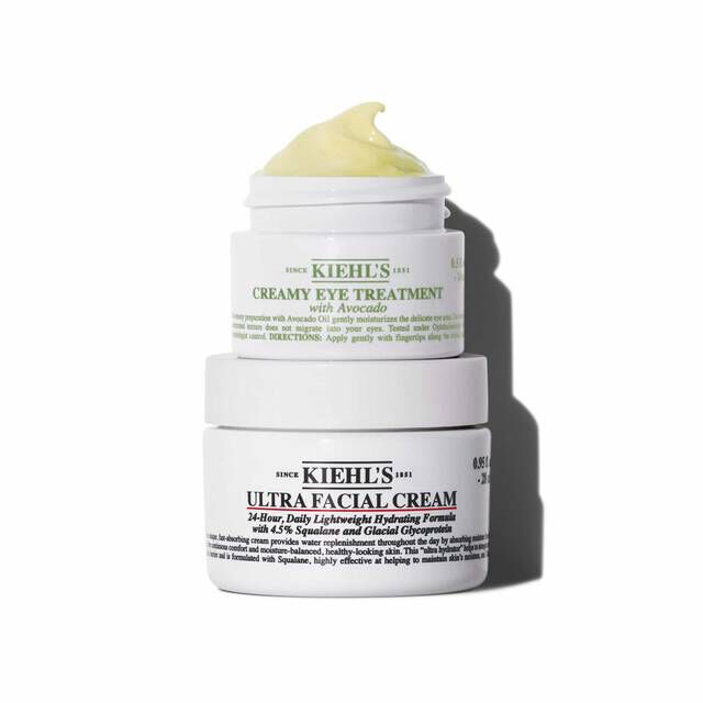 Two white jars of Kiehl's products are stacked vertically. The top jar is "KIEHL'S CREAMY EYE TREATMENT with Avocado" showing a yellow-green swirl of product on top. The bottom jar is "KIEHL'S ULTRA FACIAL CREAM". Both jars are casting shadows on a white background.