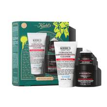 A graphic featuring a dark teal Kiehl’s gift box with white and gold accents on the left. The box contains three products. To the right of the box, the same three products are displayed outside of the box: a white tube of "KIEHL’S ULTRA FACIAL BARRIER-HYDRATING CLEANSER," a small black and red jar of "KIEHL’S AGE DEFENDER EYE REPAIR," and a larger black and red jar of "KIEHL’S AGE DEFENDER CREAM MOISTURIZER." The box is decorated with bright green graphic paint splatters and a bow design and features the text "YOUTH STILL GOT IT" at the bottom.