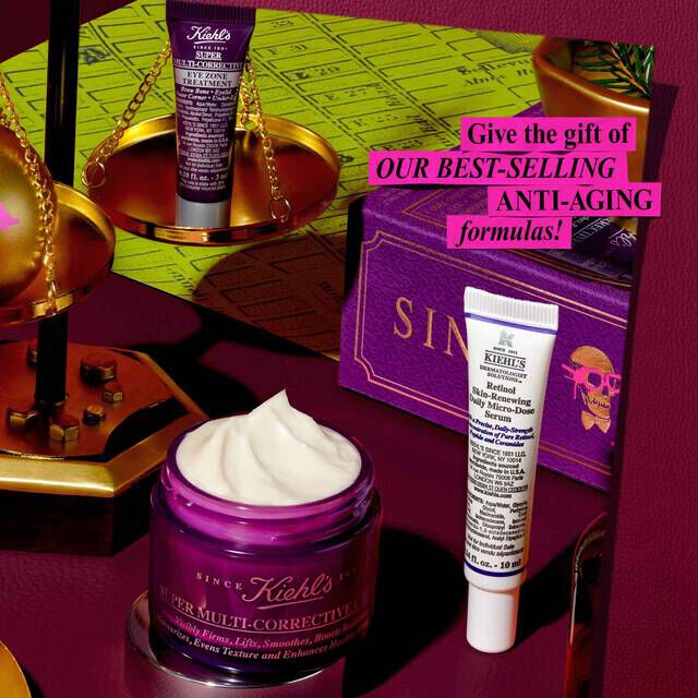 A festive graphic, set against a dark red background with a green map pattern in the back, features several Kiehl's products and holiday decorations. A small purple tube of "Kiehl's Super Multi-Corrective Eye Zone Treatment" is balanced on a gold scale. A purple jar of "Kiehl's Super Multi-Corrective Cream" with visible cream is in the foreground. A white and purple tube of "Kiehl's Retinol Skin-Renewing Daily Micro-Dose Serum" stands next to a purple gift box with "SINCE 1851" and a skull design. A pink box with white text reads "Give the gift of OUR BEST-SELLING ANTI-AGING formulas!"